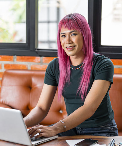 Persona con cabello rosa escribiendo en un portátil, sentada en un sofá